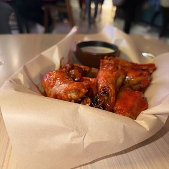 Moodswing - Da Wings - A basket of buffalo chicken wings coated in spicy sauce is placed on a wooden table. The wings are served in a paper-lined container with a small black cup of dipping sauce. The background shows a blurred restaurant setting with people standing and sitting, indicating a casual dining environment. - Venues & Event Spaces Near Me - Denver, Colorado