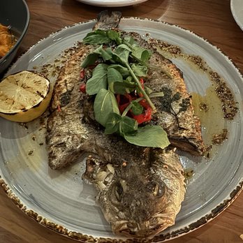 Marina - Branzino alla Griglia - A whole grilled fish with crispy skin and visible eyes is presented on a large ceramic plate. It is garnished with fresh green leaves and sliced red peppers, with a lemon half placed beside it. The dish appears to be served in a restaurant setting, with empty plates and utensils visible in the background. - Italian Near Me - Pasadena, California