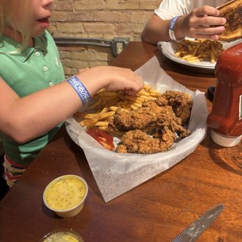 Ruby Owl - Chicken tenders - Two children are seated at a wooden table, eating fried chicken, French fries, and toast. The table has ketchup, mustard, and a serrated knife. The background features a brick wall. - Gastropubs Near Me - Oshkosh, Wisconsin