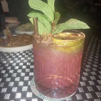 The Fed - Gerri Halliwell - A cocktail featuring a pinkish hue is presented in a glass with a salted rim. It is garnished with fresh mint leaves and a lime slice, with a straw inserted. The drink is placed on a textured, black-and-white patterned table, with some food dishes visible in the background, indicating a dining or bar setting. - Pubs Near Me - Boston, Massachusetts