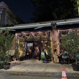 The image shows the exterior of a restaurant at night, featuring an outdoor seating area with several groups of people dining. The space is decorated with string lights hanging above, creating a warm ambiance. Potted plants are placed around the seating area, and the entrance to the restaurant is visible with a glass door. The scene includes a traffic cone on the street in the foreground and a dark sky overhead.
