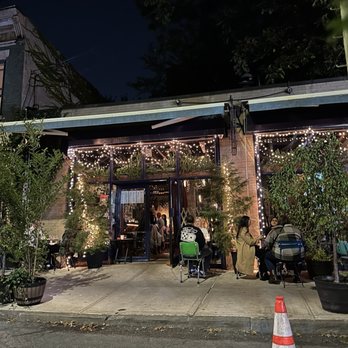 Bijan's - The image shows the exterior of a restaurant at night, featuring an outdoor seating area with several groups of people dining. The space is decorated with string lights hanging above, creating a warm ambiance. Potted plants are placed around the seating area, and the entrance to the restaurant is visible with a glass door. The scene includes a traffic cone on the street in the foreground and a dark sky overhead. - Persian/Iranian Near Me - Brooklyn, New York