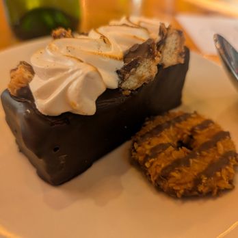 M'tucci's Italian - Girl Scout cookie themed!  House Made S'mora:  Black Velvet brownie, chocolate caramel ganache, Caramel Delight pieces! - A dessert consisting of a rectangular chocolate-covered item topped with whipped cream and cookie pieces. The dessert is presented on a white plate, with a cookie placed beside it. The background shows a wooden table and a spoon, indicating a dining setting, likely at a restaurant. The dessert appears rich and indulgent, with a combination of chocolate, whipped cream, and cookie textures. - Italian Near Me - Albuquerque, New Mexico