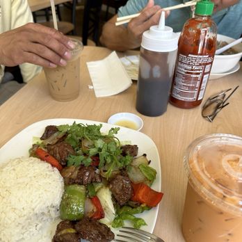 Saigon Express - Bò lúc lắc - A plate of beef stir-fry with vegetables such as bell peppers, onions, and cilantro, served with a portion of white rice, is placed on a wooden table. Two beverages are visible: a glass of iced coffee with milk and a plastic cup of iced tea or similar drink. Condiments including soy sauce and hot sauce are also on the table. The background shows a casual restaurant interior with other diners and chairs. - Vietnamese Near Me - Berkeley, California