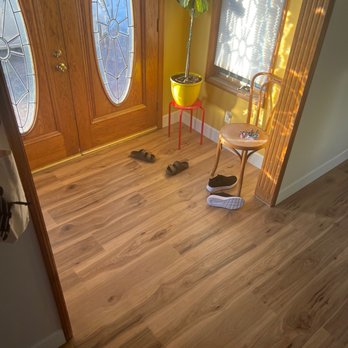 Stockdale Tile - A wooden front door with decorative glass panels, a potted plant on a red stand, and shoes scattered on the wooden floor near a window. - Carpeting Near Me - Tehachapi, California