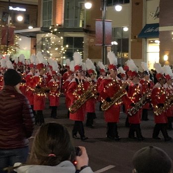 Southlands - A marching band dressed in red uniforms with white plumes on their hats performs in a street at night. The band members are playing saxophones and are arranged in rows. Spectators are visible in the foreground, some taking photos. The background includes illuminated buildings and decorative lights, indicating a festive event or holiday parade. - Shopping Centers Near Me - Aurora, Colorado