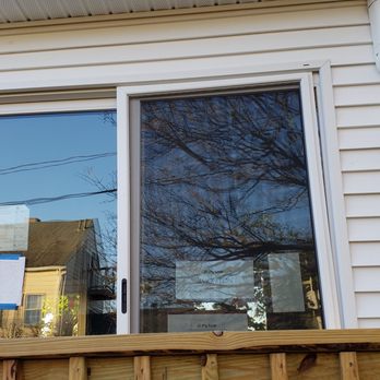 Avatar Construction - We've been having trouble with this slider since day 1.  Notice the screen. - A sliding glass door on a white house exterior, reflecting trees and neighboring houses, with two outdoor wall-mounted lights. - General Contractors Near Me - Watertown, Massachusetts