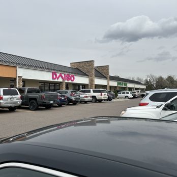 Daiso - Outside view-right next to Dollar Tree. - The image shows the exterior of a Daiso store located in a shopping plaza, with a parking lot filled with various parked cars. The storefront features a prominent pink Daiso sign, and adjacent to it is a Dollar Tree store. The sky above is overcast with gray clouds, and the area appears to be in a suburban setting with some trees visible in the background. - Discount Store Near Me - Aurora, Colorado