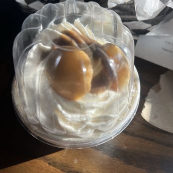 Duke City BBQ - Banana pudding - A banana pudding dessert is presented in a clear plastic cup, topped with whipped cream and banana slices. The cup is placed on a wooden surface, with some crumpled food wrappers and napkins visible in the background. The image captures the dessert's creamy texture and fresh banana toppings. - Barbeque Near Me - Albuquerque, New Mexico
