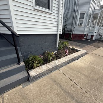 Simone & Sons Landscape Construction - After - A small garden bed with soil and plants, including some with green leaves, is bordered by gray stone bricks. Two small American flags are placed in the soil. The garden is adjacent to a house with white vinyl siding, a window, and a gray painted foundation. Concrete sidewalk and steps with a black handrail are also visible. - Landscaping Near Me