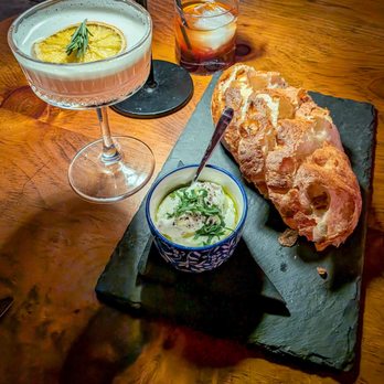 Johnny Seesaw's - Homemade Focaccia - A cocktail with lemon garnish, a bowl of creamy dip topped with herbs, and a rustic bread loaf are displayed on a dark slate serving board at a restaurant. - American Near Me - Peru, Vermont