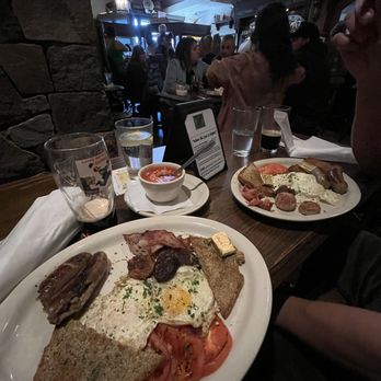 Two Fools Tavern - Irish breakfast Sunday Special - A table at Two Fools Tavern in Albuquerque, NM, displays Irish breakfast dishes with eggs, toast, sausage, bacon, and mushrooms. There are drinks including water, beer, and a glass with a straw. The background shows a busy pub interior with patrons, a stone wall, and ambient lighting, indicating a lively Irish pub atmosphere. - Irish Near Me - Albuquerque, New Mexico