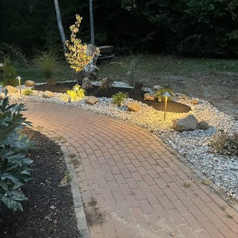Dreamscapes - a brick pathway with a stone bench in the background - Landscaping Near Me