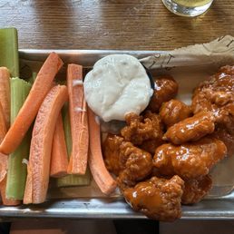 Buffalo Boneless Wings - Buffalo boneless chicken wings coated in sauce are placed on a metal tray, accompanied by celery sticks and a cup of ranch dressing. The tray is lined with paper, and the background shows a wooden table with a glass of beverage.
