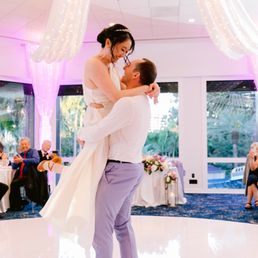 A bride and groom are dancing together at a wedding reception in a decorated indoor venue with large windows showing outdoor greenery. The bride is wearing a white dress and the groom is in a white shirt and light-colored pants. Guests are seated at tables, some clapping and watching the couple dance. The venue features draped curtains, string lights, and a chandelier, creating a festive atmosphere.