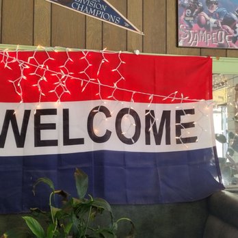Alpine Rose Cafe - a welcome sign hanging from the ceiling - A fabric banner with the word 'WELCOME' in bold black letters is hung on a wall. The banner has horizontal stripes in red at the top, white in the middle, and blue at the bottom. String lights are draped across the top of the banner. The background includes a wooden wall, framed pictures, and a window with plants visible outside. - American Near Me - Walsenburg, Colorado