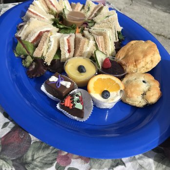 Lovey's Tea Shoppe - Queens tea service tray of sandwiches, scones, and Halloween themed petit fours with cream, curd, and jam served in plastic containers - A blue serving tray contains an assortment of finger sandwiches with various fillings, arranged in a circular pattern with greens and cucumber slices. There are two scones and several small dessert cups with fruit and chocolate decorations, including a lemon tart, an orange slice with blueberries, and small decorated chocolates. The tray is placed on a floral tablecloth, with outdoor seating visible in the background. - British Near Me - Pacifica, California