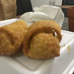 two fried food items on a plate - Two fried egg rolls are placed on a white foam plate, with a crumpled paper towel behind them. The background includes a table, chairs, and a water bottle.