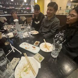 Four people are seated at a restaurant table with various empty dishes, chopsticks, glasses, and drinks, including a Pepsi can and a water bottle. The background shows a bar area with bottles and a kitchen with a vent hood. The table has remnants of a meal, and the group appears to be in conversation or waiting for the next course.