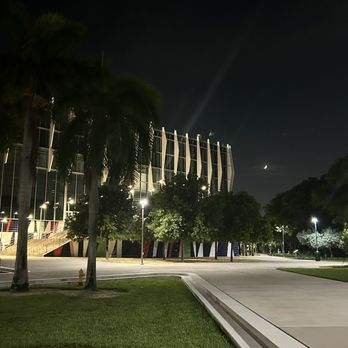 Rum Room - Exterior - Nighttime outdoor scene featuring a modern architectural building with vertical structural elements. The area is lit by streetlights, with trees and a grassy area in the foreground. The sky is dark with a visible crescent moon and a faint star. The scene appears to be in an urban or campus setting with paved walkways and minimal activity. - Cocktail Bars Near Me - Miami Beach, Florida