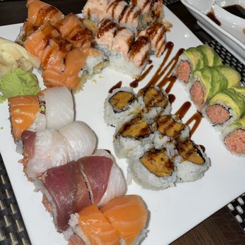 Ichiban - Ayce sushi - The image shows a white square plate filled with an assortment of sushi rolls, including salmon, tuna, and avocado-topped varieties. Some rolls are drizzled with sauces, such as eel sauce and spicy mayo, and garnished with wasabi. The sushi is arranged in groups, with a soy sauce dish visible in the background. The setting appears to be a restaurant with a dark wooden table and woven placemats. - Asian Fusion Near Me - Levittown, New York