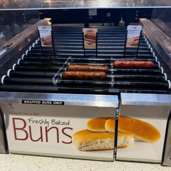 Kwik Trip - sausage - A stainless steel roller grill with several sausages cooking on it, located inside a convenience store. The grill is covered with a glass shield, and there are small signs behind the sausages indicating different hot dog options. Below the grill, a sign displays an image of buns and the text 'Open here for Freshly Baked Buns.' The setup is part of a family-owned convenience store offering ready-to-eat food items. - Gas Stations Near Me - Roberts, Wisconsin