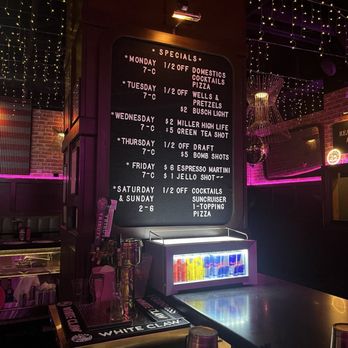 Killer Queen - The image shows the interior of a bar with a large blackboard menu listing weekly specials, including discounts on cocktails, pizza, and drinks for each day. The menu is illuminated by a small overhead light. Below the menu, a glass-front fridge contains brightly colored cans. The background features string lights and decorative lighting fixtures, creating a cozy, festive atmosphere. The bar counter has various bar tools and glasses, with a dark, modern decor style. - Pizza Near Me - Cincinnati, Ohio