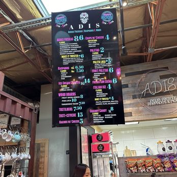 Adis - The image shows a digital menu board at Adis, a beverage and snack shop in Tucson, AZ. The menu includes aguas frescas, raspados, ice cream, wood boards, and other treats, with prices and flavor options. The background features part of the shop interior, including shelves with cups and snacks, and a woman seated below the menu. - Ice Cream & Frozen Yogurt Near Me - Tucson, Arizona