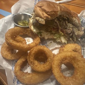 Islands Restaurant - Customized burger - A partially eaten burger with a bun, lettuce, tomato, and beef patty is placed on a tray lined with branded paper. Next to the burger are several onion rings and a small metal container with pickles. The tray is on a wooden table, with some condiments and utensils visible in the background. - Burgers Near Me - Temecula, California