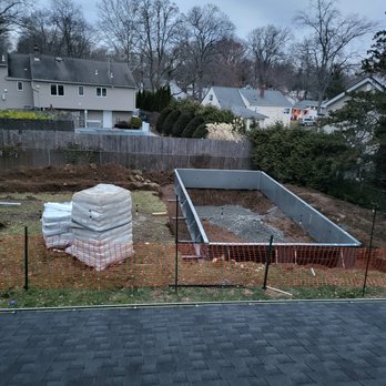 The Pool Boss - Walls constructed - A backyard scene showing a rectangular pool foundation under construction, with metal sides installed and gravel inside. Construction materials, including wrapped items and boxes, are placed nearby. The area is enclosed with orange safety fencing, and neighboring houses and trees are visible in the background. - Pool & Hot Tub Service Near Me - Wayne, New Jersey