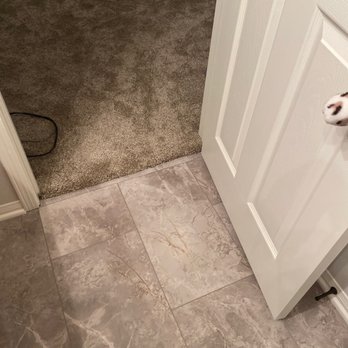 Flooring Direct Temecula - Bathroom abd bedroom - The image shows a tiled floor leading into a room with carpet, with a white door partially open, revealing part of a dog’s face. - Carpeting Near Me - Temecula, California