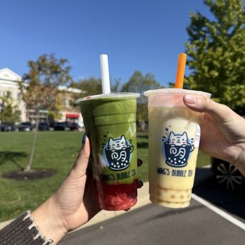 Ming’s Bubble Tea - Two bubble tea beverages are held in front of a park or outdoor area with trees and a parking lot. One drink is green with red jelly at the bottom, and the other is a creamy color with orange and pink toppings. Both cups have the Ming's Bubble Tea logo and are topped with plastic lids and straws. The background features a clear blue sky, green trees, and a building with parked cars. - Bubble Tea Near Me - Hudson, Ohio