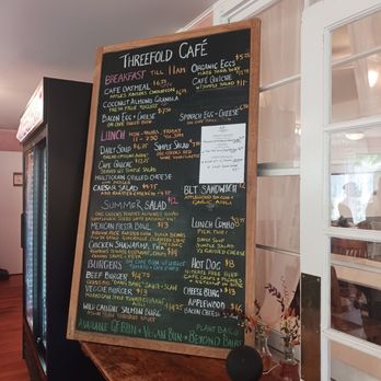 Threefold Cafe - Daily menu - The image shows a blackboard menu at Threefold Café, featuring sections for breakfast, lunch, and BLT sandwiches. The menu includes items like oatmeal, coconut almond granola, bacon egg & cheese, and various salads and burgers. Prices are listed next to each item, with some vegan and gluten-free options highlighted. The setting appears to be inside the café, with a wooden counter and a glass display case nearby. - Coffee & Tea Near Me - Chestnut Ridge, New York