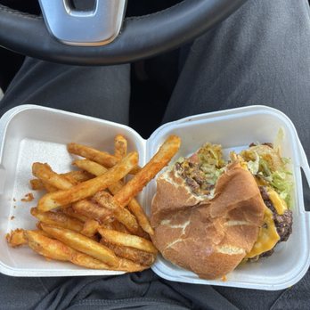 Rex's Hamburgers - The image shows a foam takeout container with French fries and a cheeseburger, which includes lettuce, cheese, and a beef patty, held on a person's lap inside a vehicle. The person is wearing dark pants, and the steering wheel is visible in the background, indicating they are seated in the driver's seat. - Burgers Near Me - Albuquerque, New Mexico