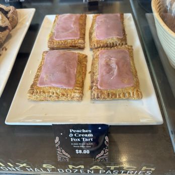Corvus Coffee Roasters - Homemade pop tarts! - Four rectangular pastries with ham slices on top are arranged on a white plate in a bakery display case, labeled as Peaches & Cream Fox Tart. - Coffee & Tea Near Me - Arvada, Colorado