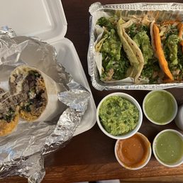 Carne asada burrito and chicken tacos - The photo features a carne asada burrito sliced in half, revealing rice, beans, and meat inside aluminum foil. Next to it is a tray with various tacos topped with guacamole, shredded lettuce, and sliced lime wedges. Four small cups contain guacamole, green salsa, and red salsa, arranged on a wooden surface. The meal appears to be from a Mexican eatery offering traditional dishes and fresh ingredients.