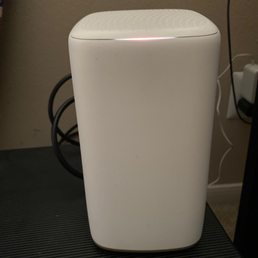 The modem. Just one cord to plug in, another to connect to cable outlet - simple! How times have changed -and I'm here for it.! - A white, rectangular electronic device with rounded edges, possibly a Wi-Fi router or modem, sits on a black textured surface in a room.