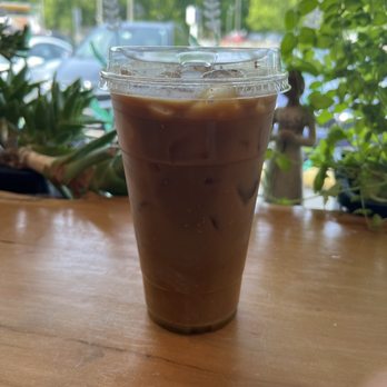 Terra Cafe - Iced Pistachio Latte - A clear plastic cup with iced coffee, containing ice cubes, is placed on a wooden table inside a cafe with greenery visible outside. - Coffee & Tea Near Me - Monroe, Connecticut