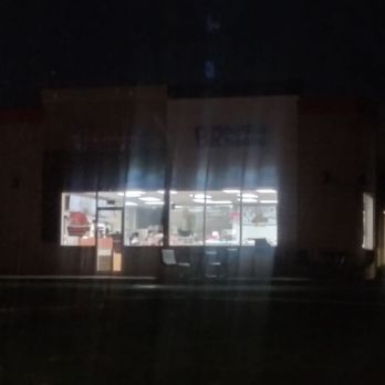Dunkin' - Coffee all night. - The image shows the interior of a fast food restaurant illuminated with bright ceiling lights, visible through a large glass window at night. The interior includes seating arrangements and counters, with some equipment and decorations visible inside. The exterior is dark, with the focus on the brightly lit interior space. - Coffee & Tea Near Me - Saint John, Indiana