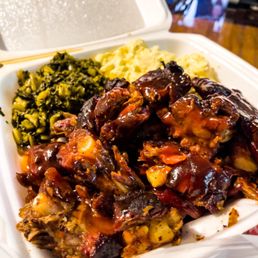 Rib Tips - A foam takeout container holds barbecue ribs with sauce, mashed potatoes, and cooked greens, likely from Pop's BBQ in Milwaukee, WI. The ribs are glazed with sauce, and the sides include mashed potatoes and greens. The container is placed on a surface with a background showing part of a restaurant or food truck setting.