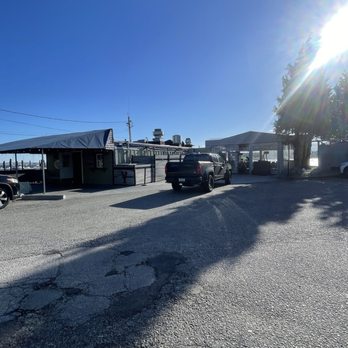 Quito's - Front entrance - A parking lot with a building structure on the left and a black pickup truck parked in the center. The lot has visible cracks and uneven pavement. The building appears to be part of a waterfront restaurant or similar establishment, with outdoor seating areas and a canopy. The sky is clear and blue, with the sun shining brightly from the right side, creating lens flare and casting shadows. Utility poles and wires are visible in the background, indicating an outdoor setting near water. - Seafood Near Me - Bristol, Rhode Island