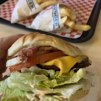 Apollo Burger - Apollo Burger and fries - A cheeseburger with lettuce, tomato, bacon, and melted cheese is held in hand, with French fries in paper on a tray in the background. - Burgers Near Me - Lehi, Utah