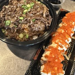 A black bowl contains sliced beef garnished with chopped green onions and sesame seeds, accompanied by a sushi roll topped with orange fish roe on a black tray.