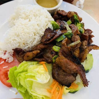 Pho 102 - A plate of grilled meat topped with chopped green onions, served alongside white rice, fresh lettuce, tomato slices, and shredded carrots. A small cup of sauce is placed above the rice. The setting suggests a Vietnamese restaurant, with condiments and beverages visible in the background. - Vietnamese Near Me - Chantilly, Virginia
