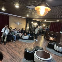 The amazing friends and staff - People are seated and standing in a lounge with dark walls, wooden floors, and modern seating, including ottomans and circular rugs.