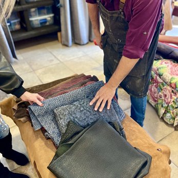 J And M Custom Boots - Choosing different types of leathers - A person wearing a dark apron and a maroon shirt is examining a selection of leather swatches and samples laid out on a table. The samples include different textures and colors, with some resembling exotic or patterned leather. Another individual is pointing at the samples, indicating interest or discussion. The setting appears to be a workshop or store specializing in leather goods, with shelves and fabric visible in the background. - Customized Merchandise Near Me - Santa Fe, New Mexico