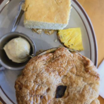 Christy Mae's Restaurant - Emily Lynn's Turkey and green chili Pot Pie with corn bread. - A breakfast plate includes a large fried chicken biscuit, a slice of pineapple with black seeds, a piece of cornbread, and a scoop of butter in a small dish. The food is arranged on a round metal tray with a fork nearby, set on a wooden table. - Sandwiches Near Me - Albuquerque, New Mexico