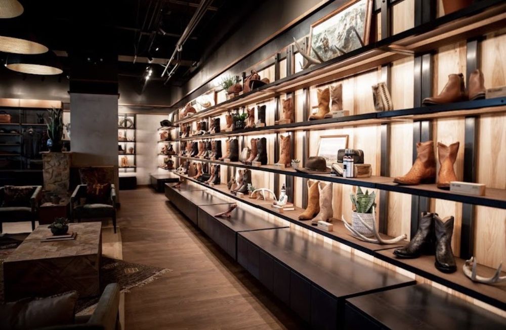 This image showcases a high-end retail store interior with a focus on western-style boots and accessories. The store features sleek, modern shelving with built-in lighting, which elegantly displays a variety of leather boots, hats, and small accessories. The warm wood tones and dark metal shelving create a sophisticated yet rustic aesthetic, aligning with a premium western brand like Tecovas.