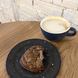 Sea Salt Caramel - A chocolate chip cookie on a black plate with white speckles and a latte in a blue mug are placed on a wooden table near a white brick wall.