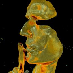 The salesman - A detailed glass sculpture shows a person's face in profile, with features such as the nose, lips, and chin clearly defined. The figure has a finger held up to the lips in a gesture of silence. The sculpture is made of transparent and colored glass, with orange and yellow hues, and is set against a black background, highlighting its intricate craftsmanship.